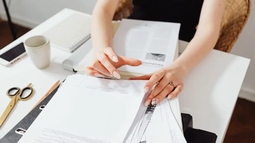 Woman organizing paperwork and documents at a desk, reviewing printed pages with manicured nails and coffee cup nearby.